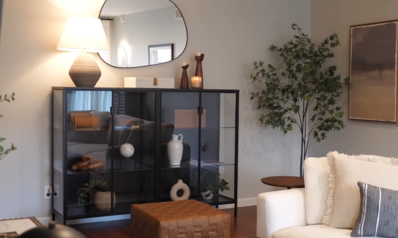 A stylish corner of a room featuring a black glass display cabinet filled with decorative objects. On top of the cabinet sits a table lamp, a box, two wooden candle holders, and an irregular shaped wall mirror. To the right is a tall artificial tree and a white sofa with a textured pillow. A brown woven leather pouf rests on the hardwood floor in front of the cabinet.