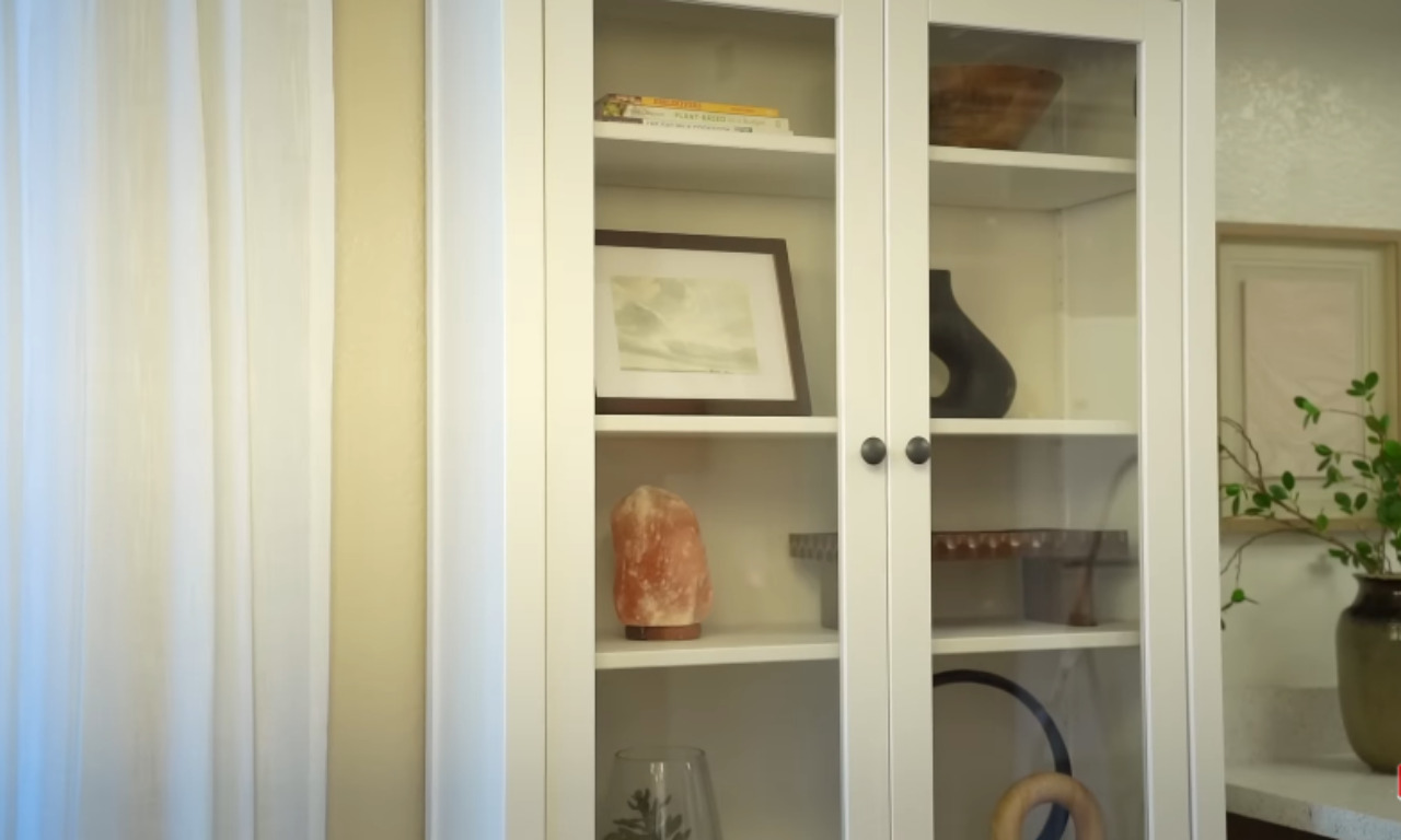 A close-up view of the white display cabinet with glass doors. The shelves inside are decorated with various items, including a framed picture, a book, a wooden bowl, a black vase, a piece of pink Himalayan salt, and other decorative objects. Sheer white curtains are visible to the left.