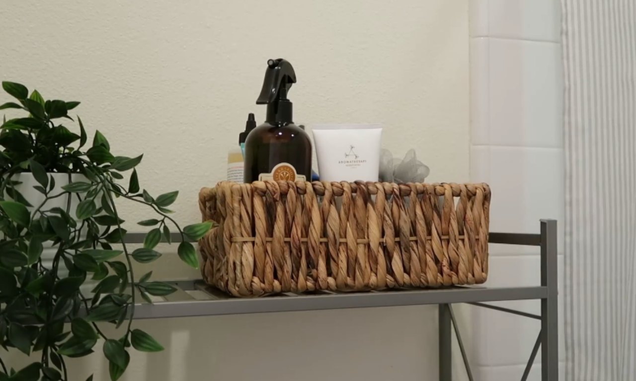 A close-up shot of a small, woven natural fiber basket resting on a metal shelving unit. The basket holds various toiletries, including a dark spray bottle and a white lotion tube labeled "Aromatherapy." To the left, a vibrant green potted plant trails downwards, providing a natural element against the light-colored wall.