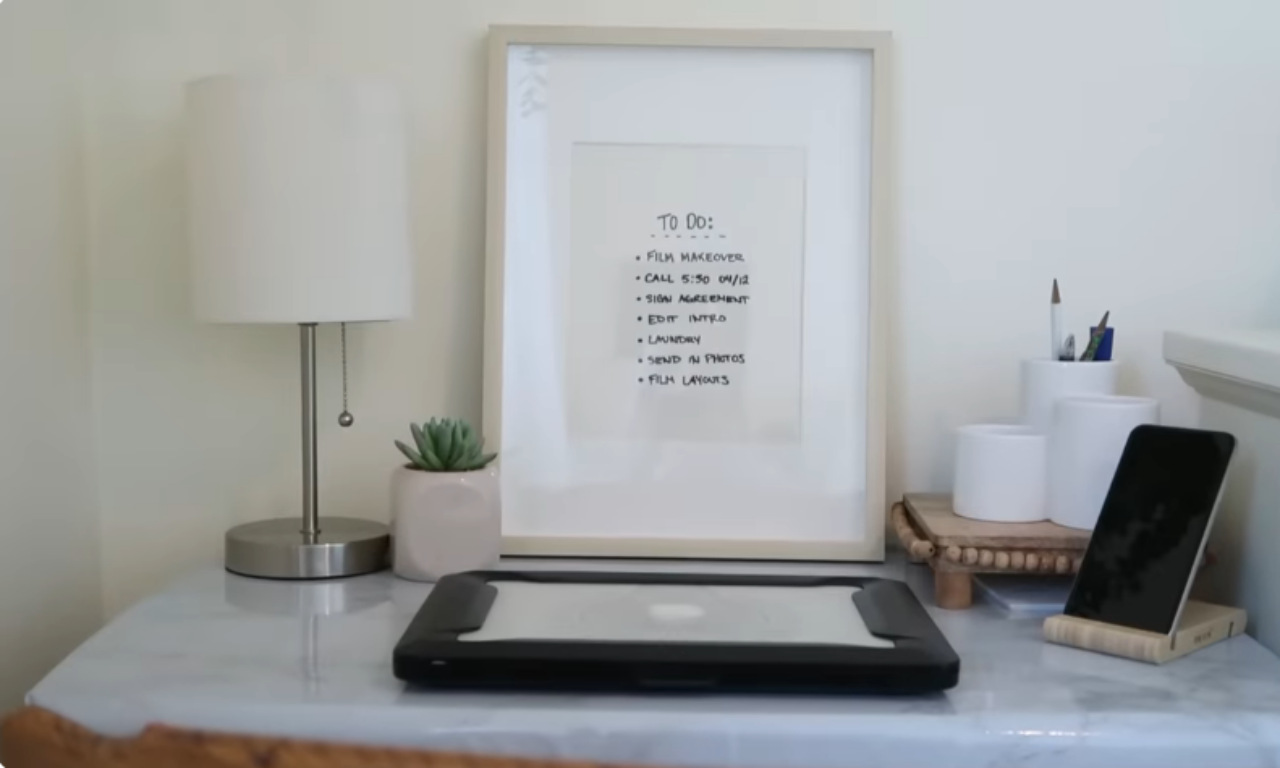 A close-up of a small, well-organized desk setup with a marble-look surface. On the desk are a silver-based table lamp with a white shade, a small potted succulent, and a framed "TO DO" list handwritten on a piece of paper. To the right are white ceramic pen holders on a small, beaded wooden stand, and a tablet or e-reader device is propped up on a wooden stand.