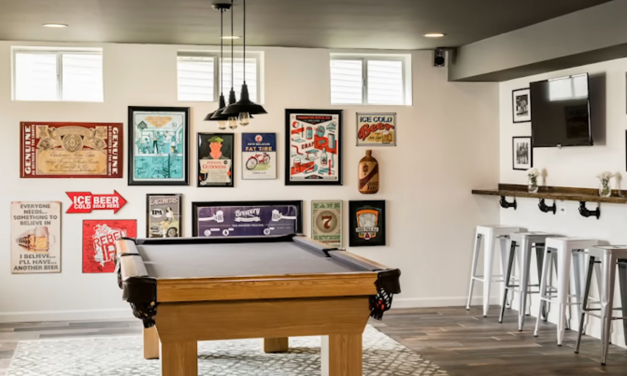 A bright, modern basement recreation room featuring a wooden pool table on a patterned rug. The white walls are decorated with a large gallery wall of vintage-style beer and brewery advertisements and signs. To the right, there is a narrow bar counter with several white metal stools.