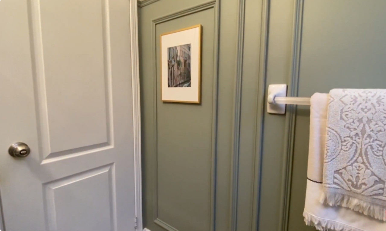 A close-up view of a bathroom wall painted a soft sage green, featuring decorative wall molding creating rectangular panels. A small framed picture with a gold-toned frame hangs in one of the panels. A white towel rack with a patterned white towel hangs to the right. The white door is visible on the left.