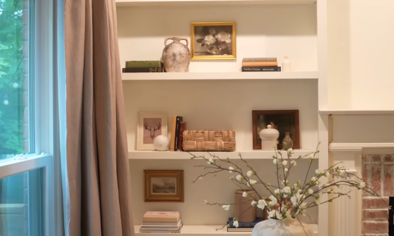 A close-up of three shelves on a white built-in bookcase styled with various decorative items, including books, small framed art, ceramic vases, and a woven basket. A light-colored curtain and window are visible on the left, and a white vase with flowering branches is in the foreground.