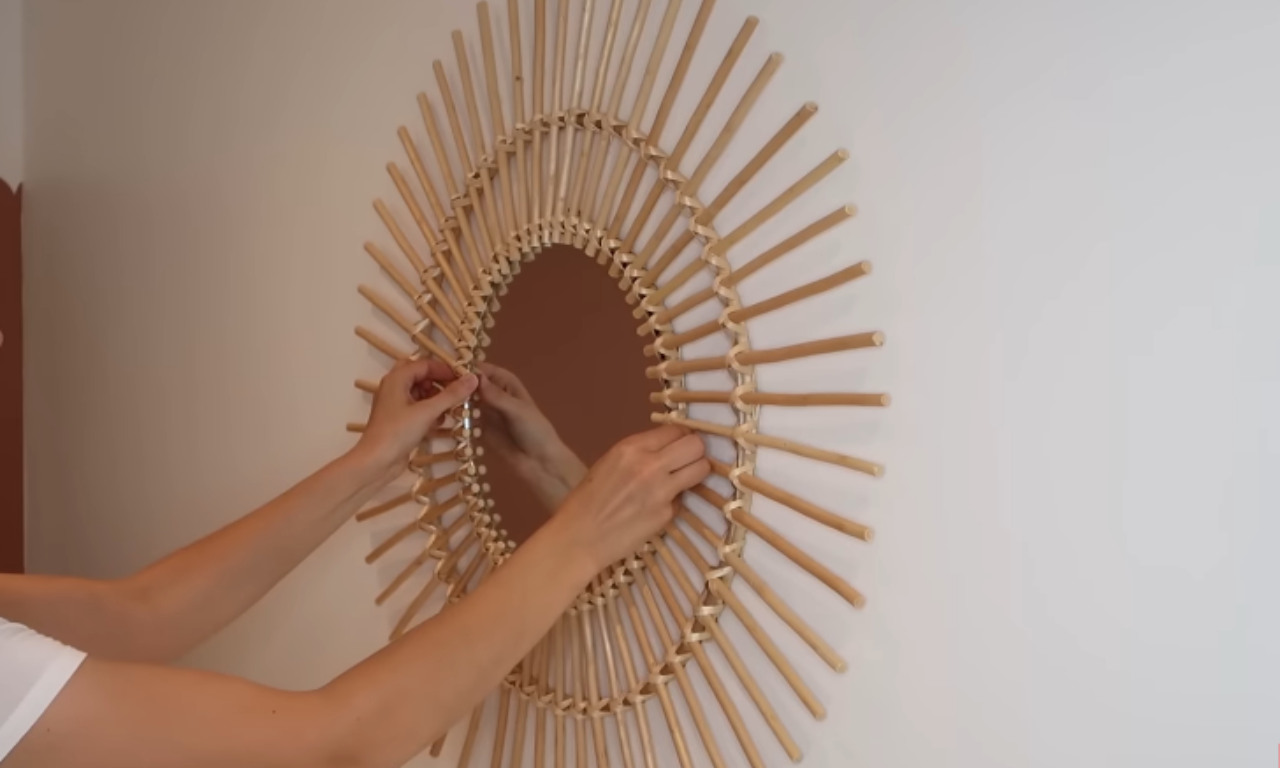 A pair of hands is shown installing or adjusting a large, round sunburst-style mirror made of natural light-colored wood or rattan on a plain white wall.