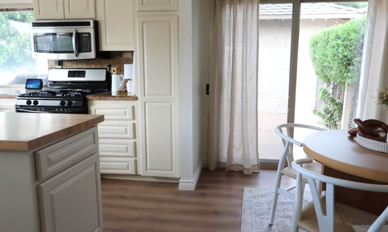 A view of a kitchen with light cream cabinets, a microwave over a stove, and a wood-look countertop. To the right is a dining area with a round table and white chairs next to a glass sliding door with sheer curtains.