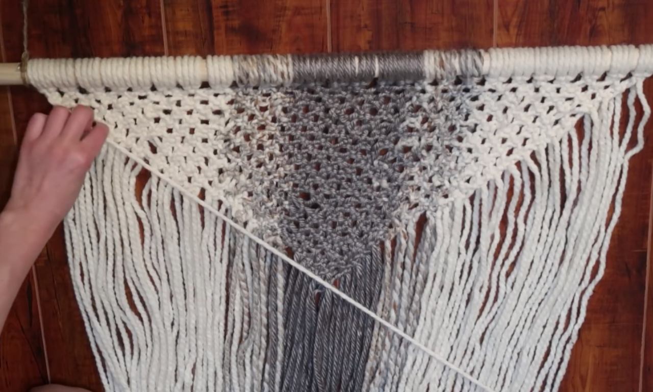 A close-up view of a macrame project in progress. A person's hand is shown attaching a cream colored cord to the wooden dowel using a Lark's Head Knot next to the existing woven section, which is an inverted triangle pattern in cream and gray. The dowel is already covered with cords.