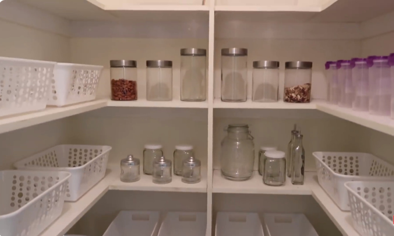 A clean, organized pantry with white shelving. The shelves are mostly stocked with clear glass jars of various sizes holding dry goods like beans and nuts, and white plastic storage baskets.