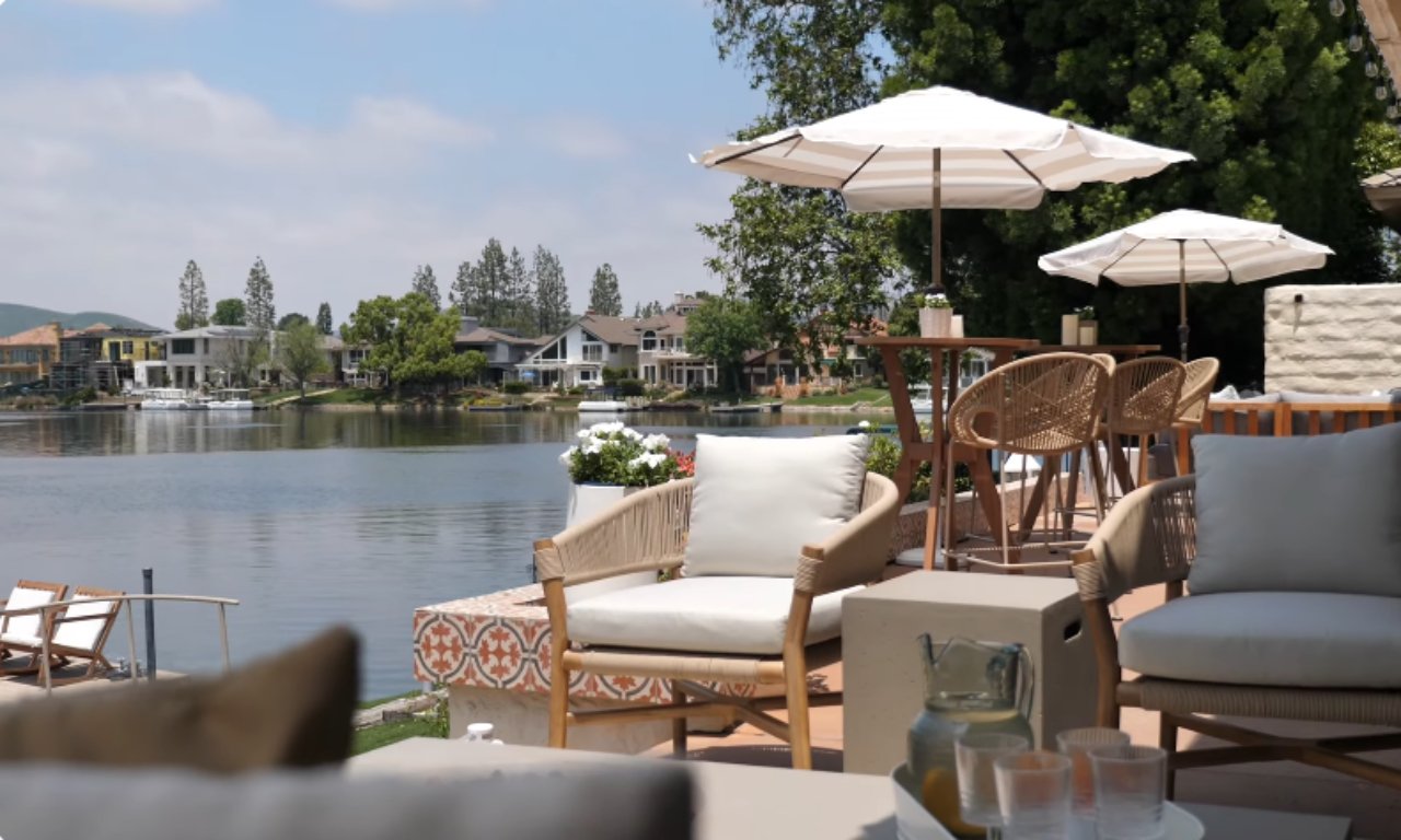 A sunny lakeside patio scene showing an outdoor living area. The foreground features a light-colored woven armchair with white cushions, a small concrete side table, and a tray with a pitcher and glasses. Beyond the seating area, there is a bar-height table with wicker stools and a striped umbrella. The background shows a large lake surrounded by houses and trees under a blue sky.