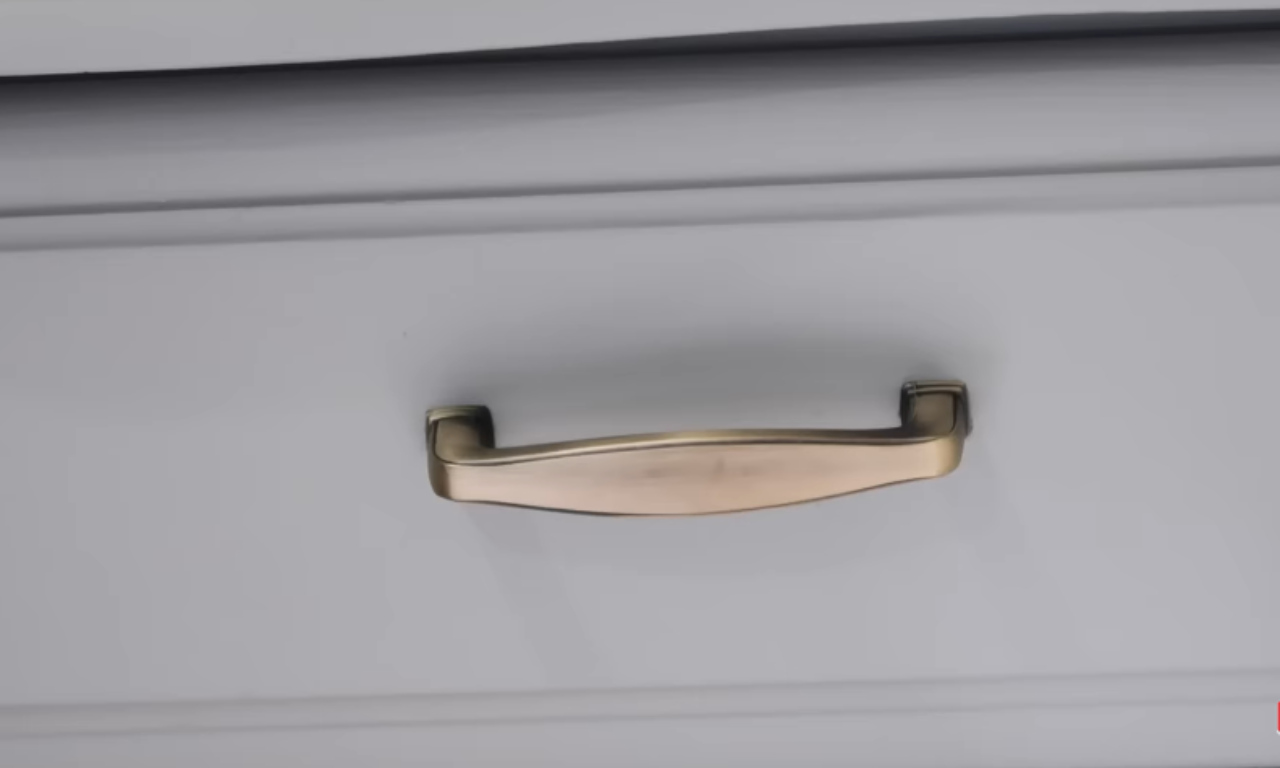 A close-up shot of a new, curved brass-toned drawer pull handle against the smooth, light gray-blue painted surface of a dresser drawer, highlighting the final detail of the furniture makeover.