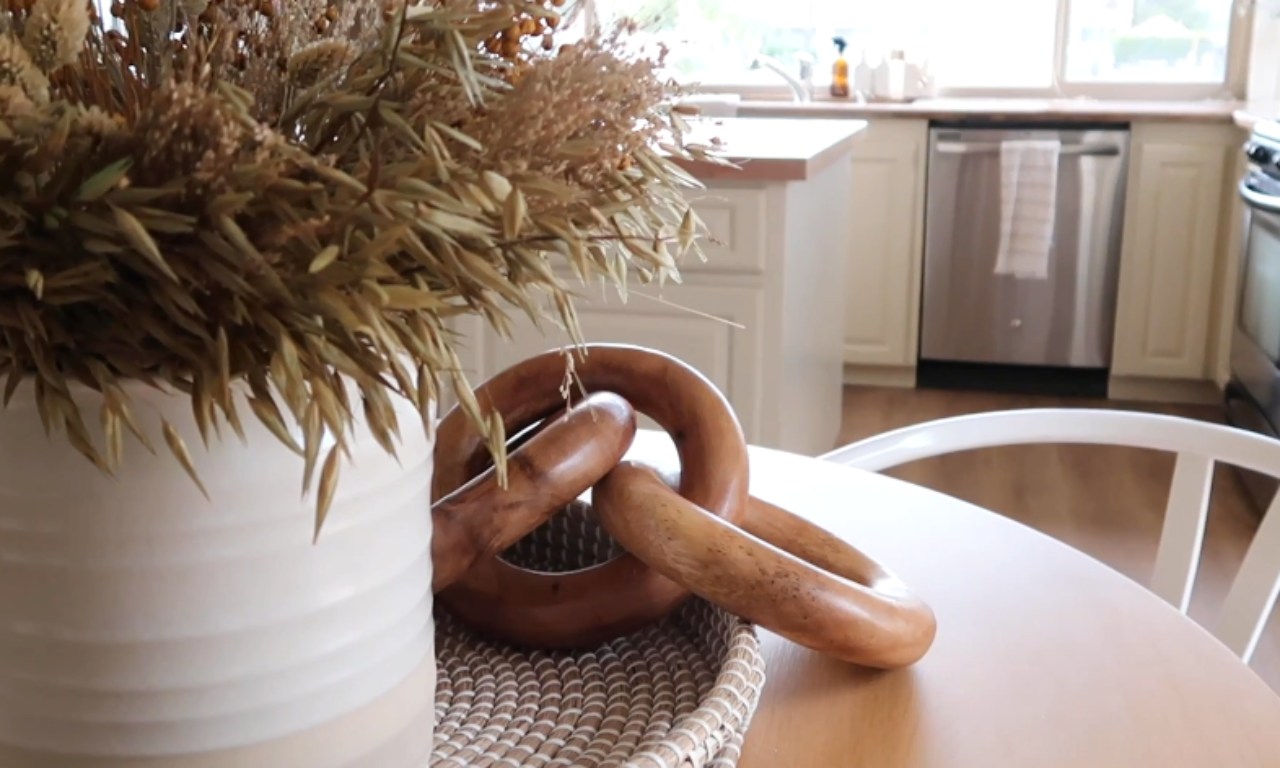 A close-up view of kitchen table decor, featuring a cream-colored vase with dried grasses and a natural wooden chain resting on a woven tray. A neutral-toned kitchen with white cabinets is softly blurred in the background.