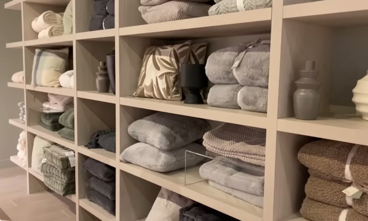 Close-up of open shelving units filled with stacked blankets, pillows, and towels in shades of grey, beige, and sage green, suggesting a section for bedding and linens in a home goods or decor store.