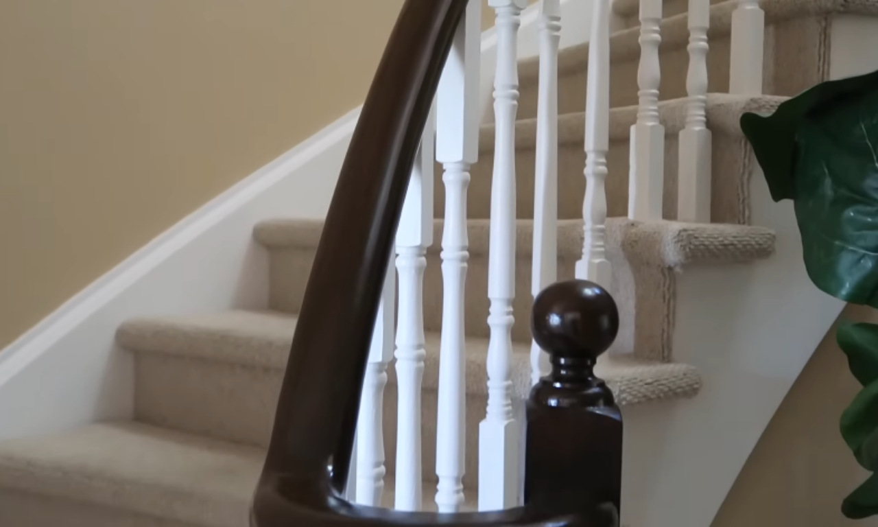 A close-up view focusing on the bottom of the staircase railing. The dark, glossy curved handrail and a dark wooden newel post cap are prominent, set against the row of white painted spindles and the white stringer (trim along the side). The stairs have a light, textured beige carpet runner.