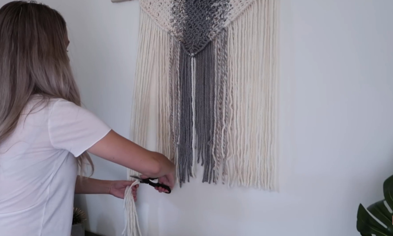 A person with blonde hair wearing a white shirt is using small scissors to trim the long, hanging strands of yarn/cord on a nearly finished macrame wall hanging. The macrame is cream and gray, and is hanging on a white wall.