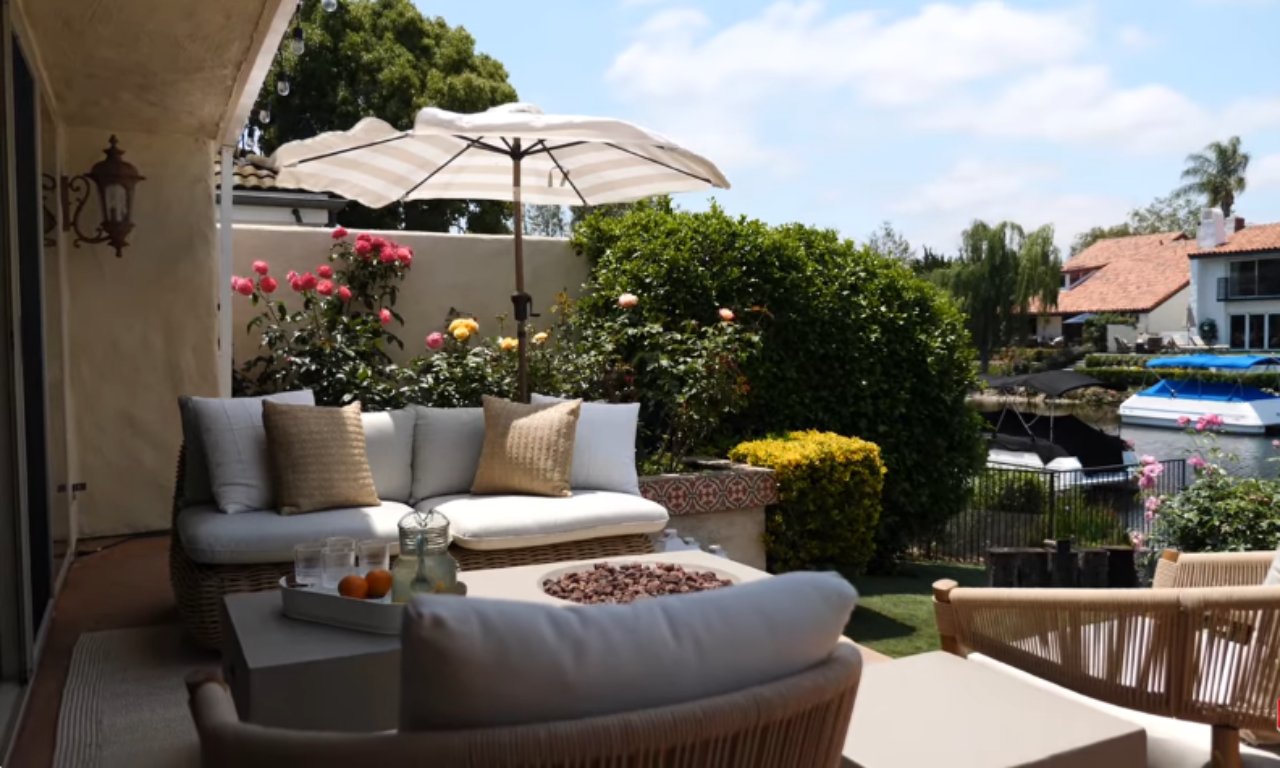 A bright outdoor patio featuring a comfortable seating area next to a lush garden. A white cushioned outdoor sofa with neutral throw pillows is positioned near a low coffee table holding a tray with drinks and a bowl of decorative pebbles. The area is partially shaded by a striped umbrella. Blooming roses and green bushes line the background, with a view of a canal and houses.