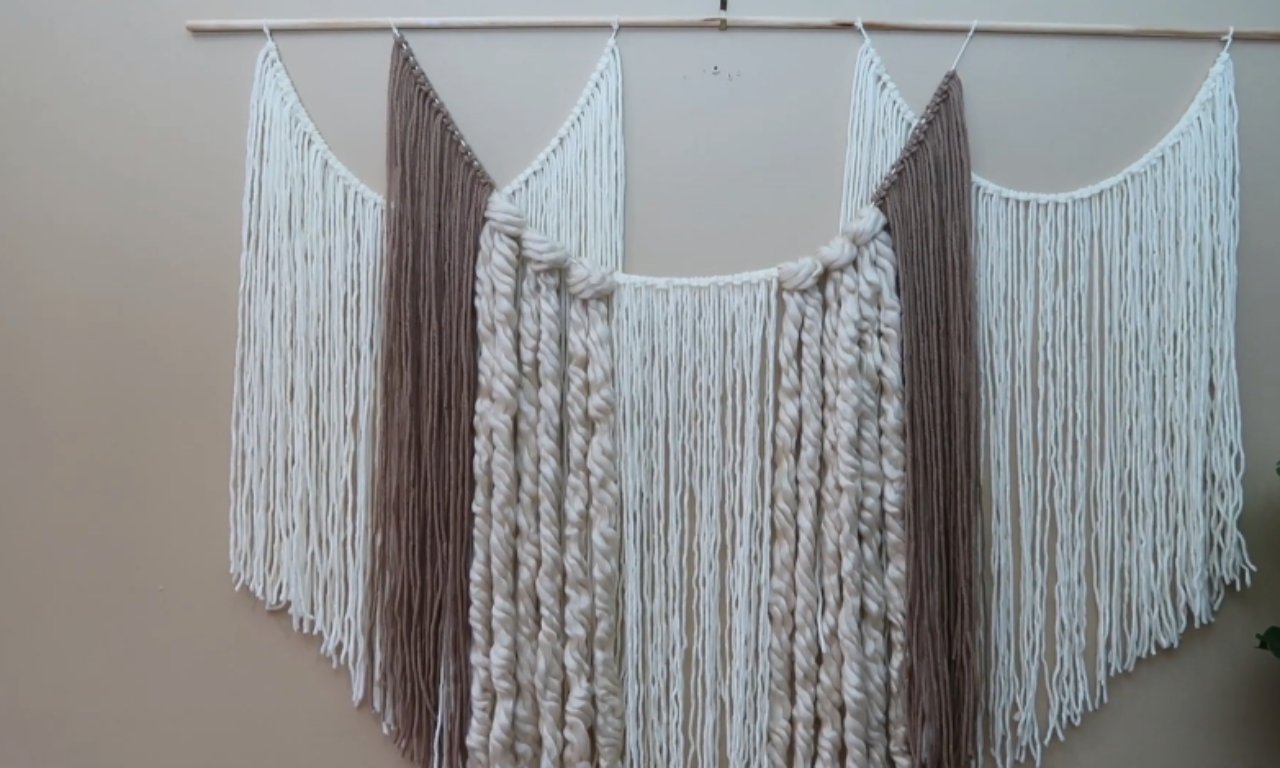 A large DIY yarn wall hanging on a light beige wall. It features an arched design, hung from a wooden dowel. The wall hanging is made of various textures of yarn in cream/off-white and brown. The center section uses thick, loosely twisted yarn in cream, flanked by straight-hanging, thinner yarns in brown and cream creating a cascading effect.