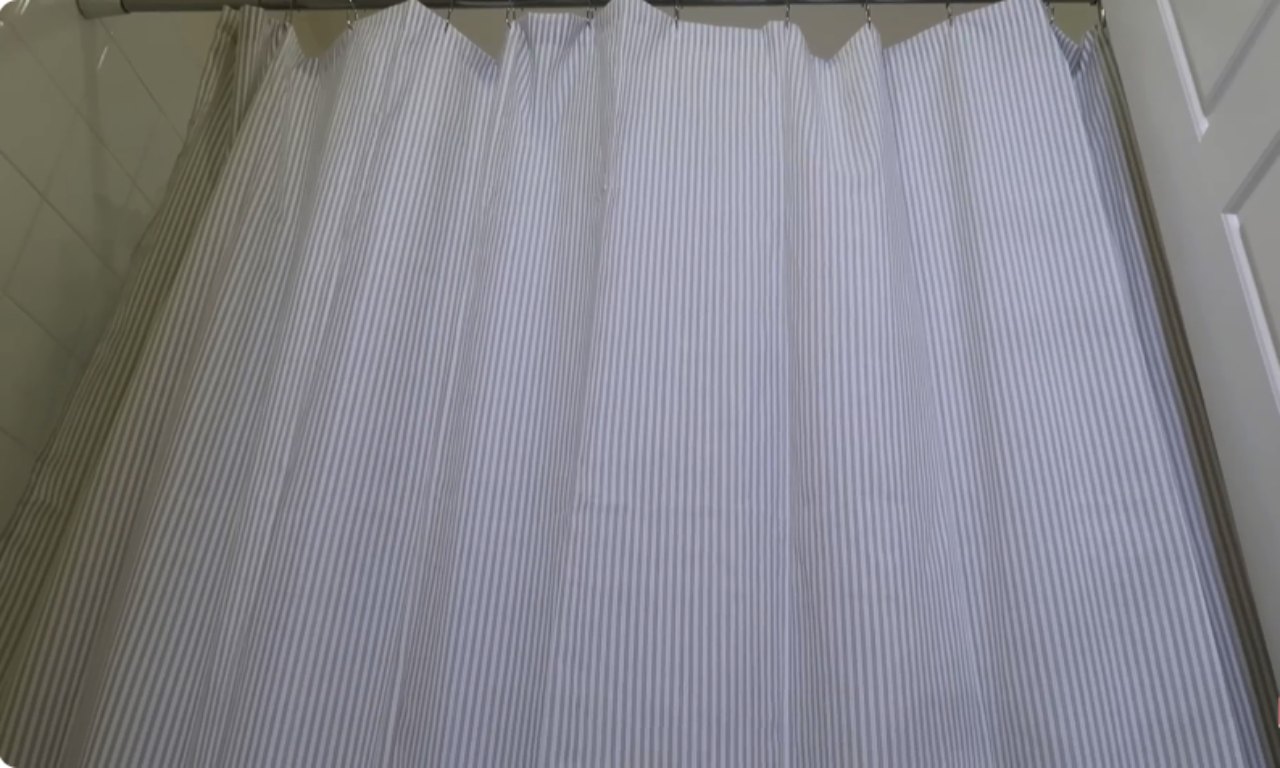 a fabric shower curtain hanging from a rod. The curtain has a classic, light grey and white vertical stripe pattern and is fully extended, obscuring the shower area.