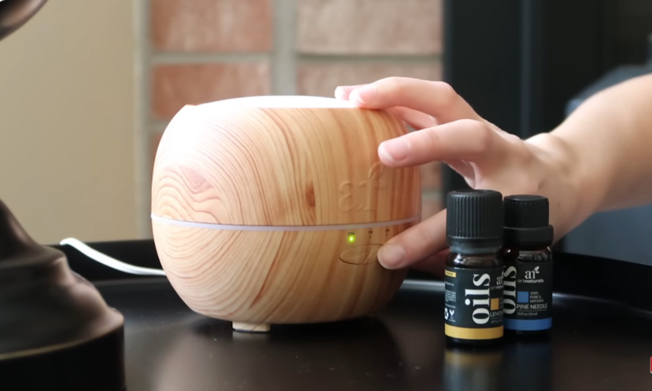 A person's hand pressing a button on a light wood-grain essential oil diffuser. Two small bottles of essential oils, one labeled "LEMON" and the other "PINE NEEDLE," are visible next to the diffuser.