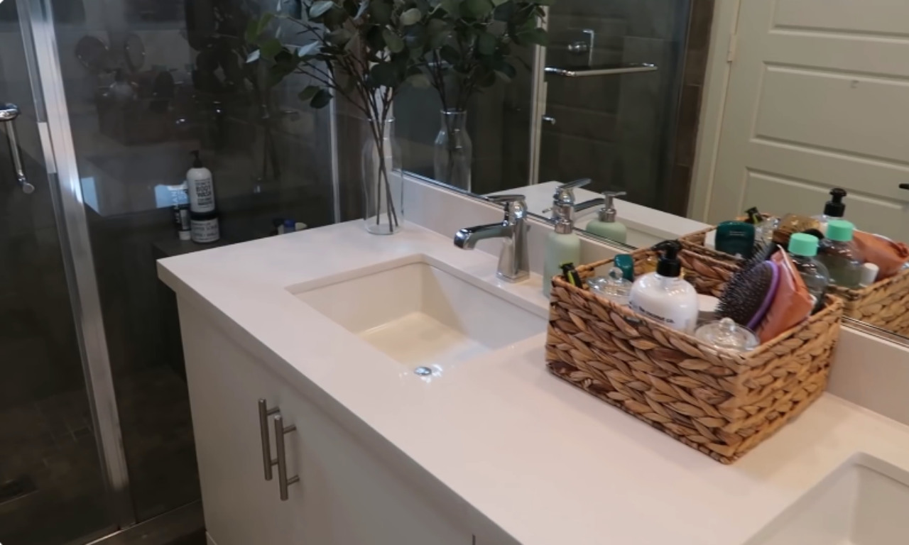 A bright, modern bathroom vanity with a white countertop and a rectangular sink. A woven basket holds various bath products and brushes next to a chrome faucet. A shower enclosure with a glass door is visible to the left.