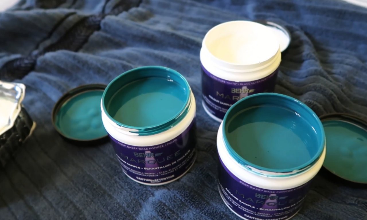Three small, open paint sample containers, two filled with a deep teal/turquoise color and one with white, sitting on a dark blue, textured fabric. The containers are labeled BEHR MARQUEE.