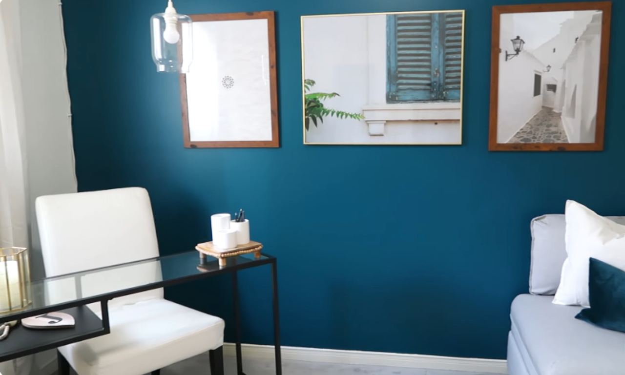 A corner of a room featuring a small desk area against a deep teal accent wall. A white chair is pulled up to a modern glass-top desk with a black frame. Various desk accessories and decorative items are visible on the desk. The wall is decorated with three framed art prints, one of which features a blue-shuttered window. A light fixture hangs from the ceiling in the upper left corner. A grey upholstered piece of furniture is partially visible on the right.