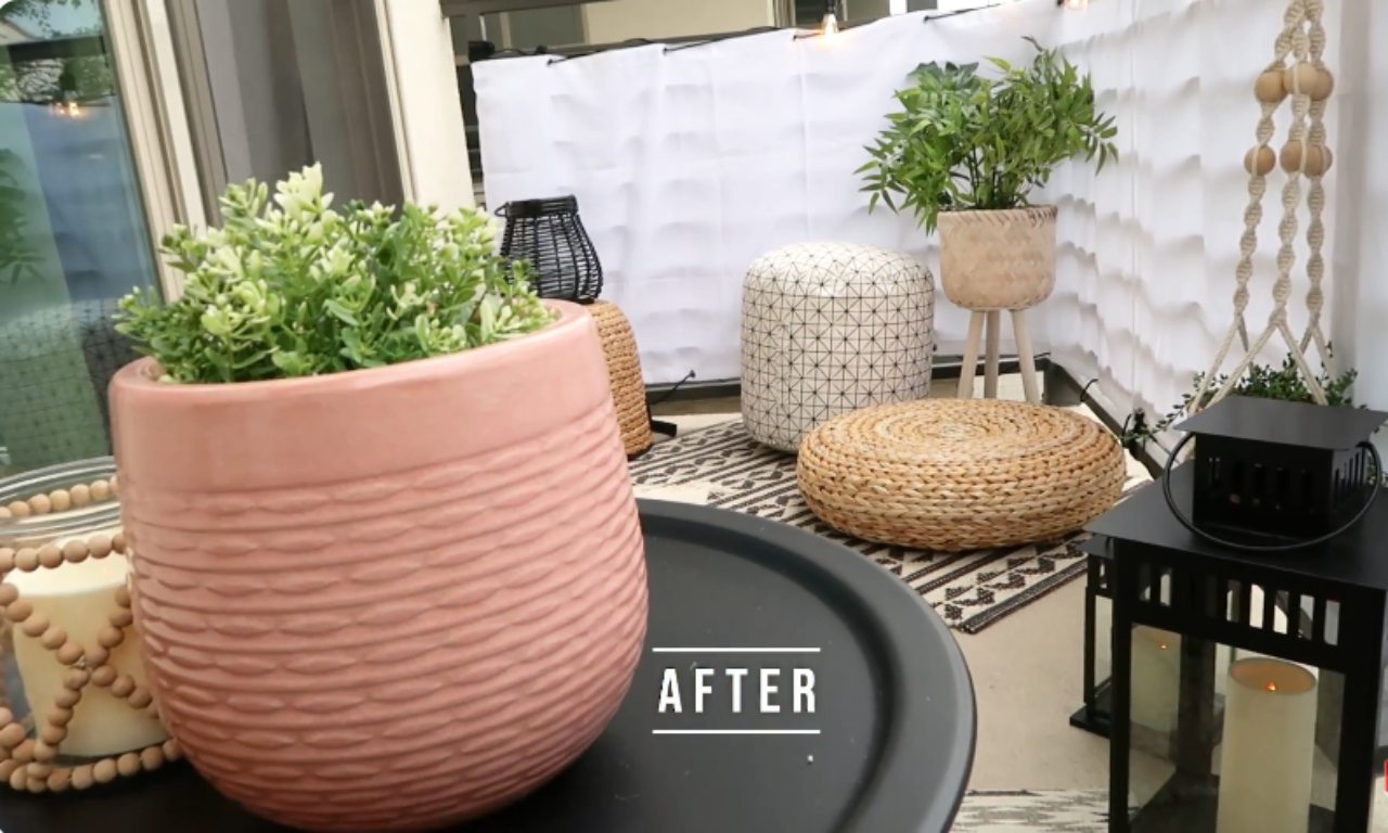 A close-up view of an "AFTER" shot of a finished balcony makeover, emphasizing a textured pink planter with a small green plant. The background shows two woven poufs and one patterned pouf on a patterned rug, a potted plant on a wooden stand, and a black lantern, highlighting the cozy, finished decor.