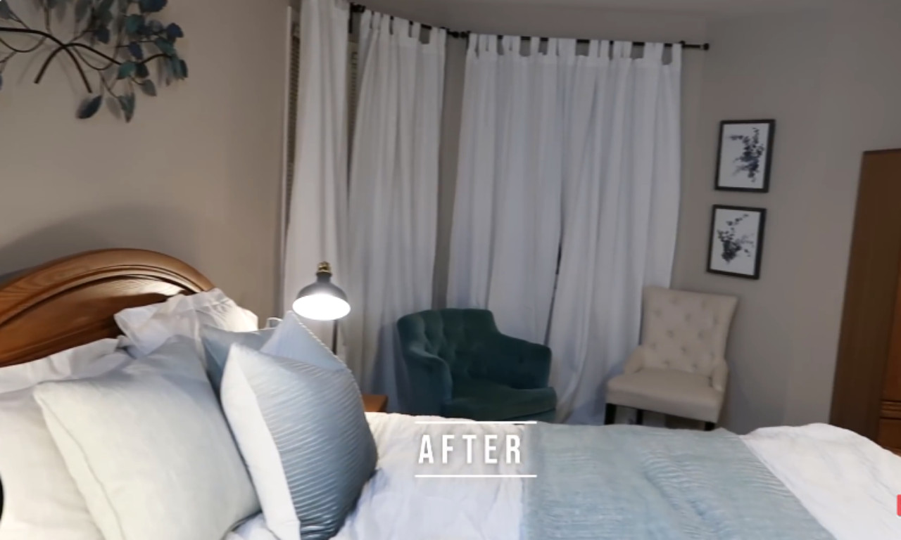 A cozy bedroom corner after a furniture rearrangement, showing a wooden bed frame, white and light blue bedding, a small lamp, and two upholstered accent chairs (one teal, one white tufted) in front of white curtains.