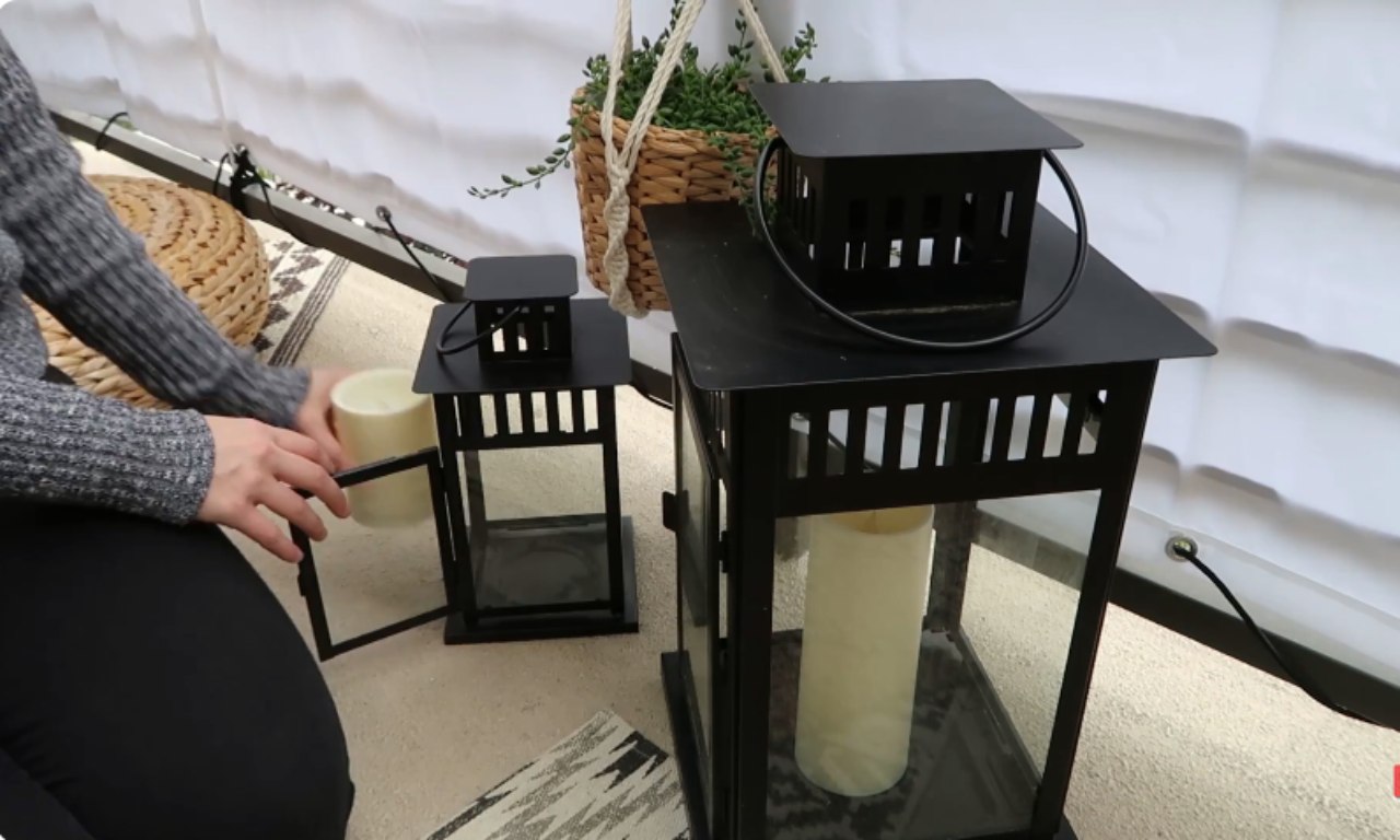 A close-up shot showing a person's hands placing a tall, cylindrical LED candle inside a large, black, square metal lantern with glass panels. A smaller, matching lantern and a hanging woven planter are visible in the background on a patterned rug.