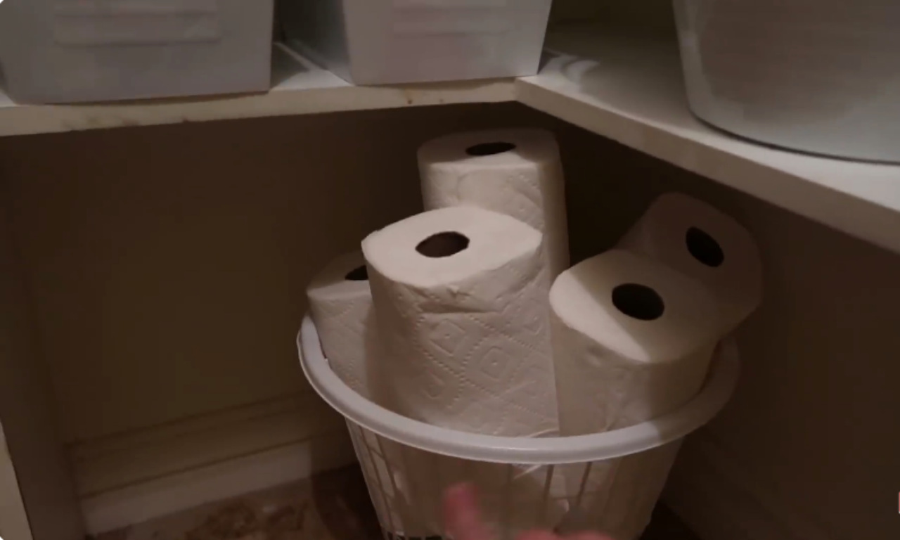 A white plastic basket on a pantry shelf filled with several rolls of toilet paper. Other white storage containers are visible on the shelves above.
