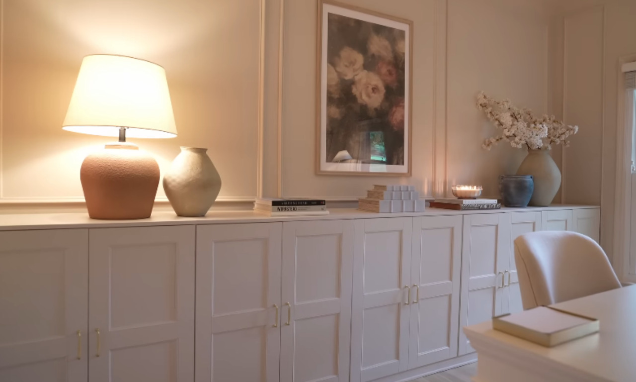 Close-up of white, paneled built-in cabinets and a shelf top in a home office. The walls and trim are painted a matching neutral color. The shelf holds an orange-brown lamp, a cream vase, stacks of books, a candle, and two large ceramic vases with white floral branches. A framed floral print hangs above the cabinets.