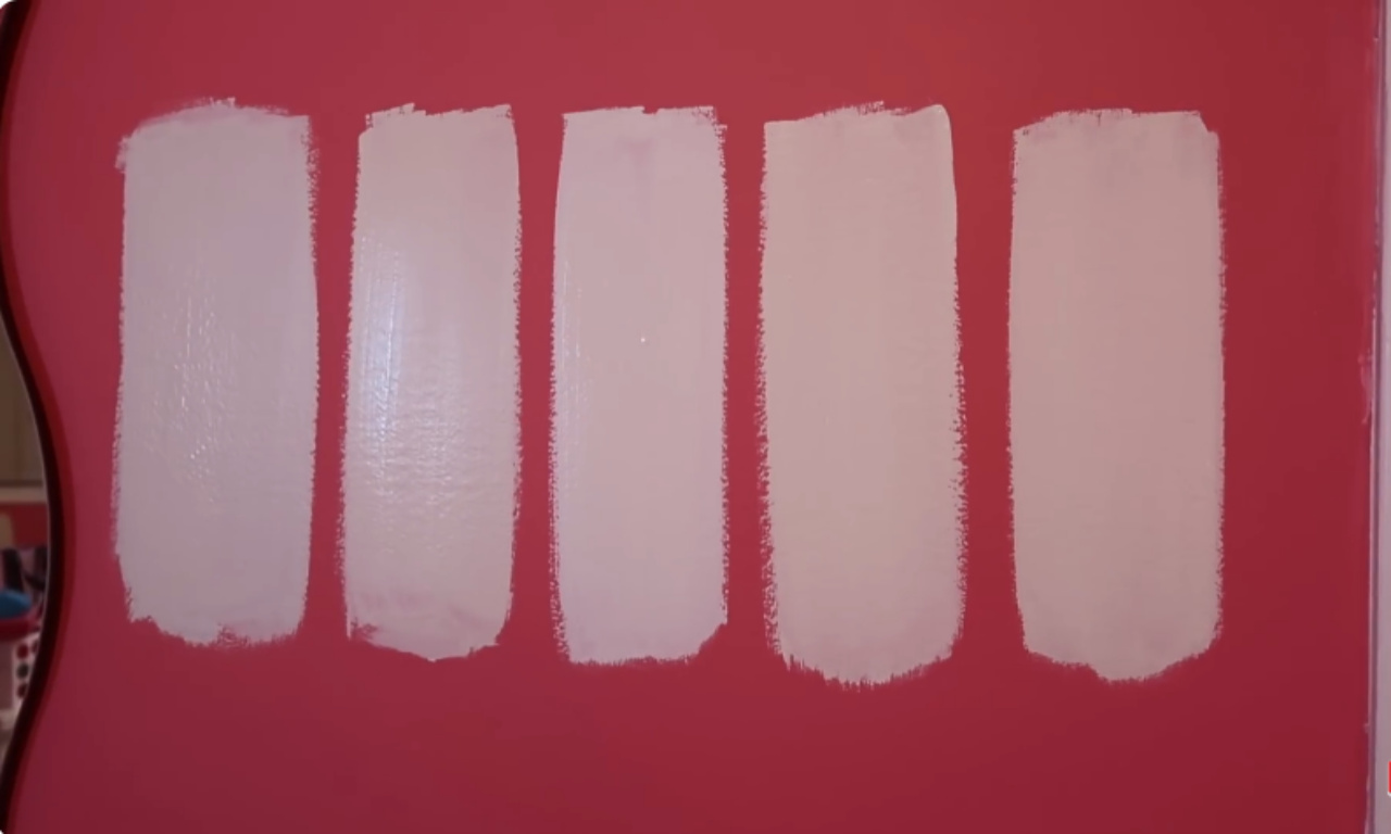 Five vertical swatches of a very light, almost white or pale pink paint color are painted side-by-side on a bright, magenta-pink wall. This shows the process of testing different shades before committing to a whole room color.