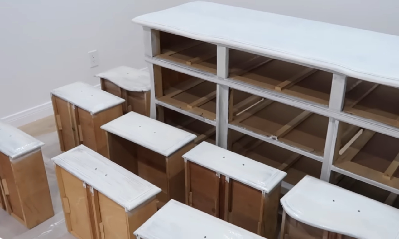 An in-progress furniture makeover shot showing the main body of a large dresser and all its detached drawers. The surfaces that will be painted are coated in a white primer, while the sides of the drawers and the interior wood are still the original light brown color. The pieces are laid out on a drop cloth in a white room.