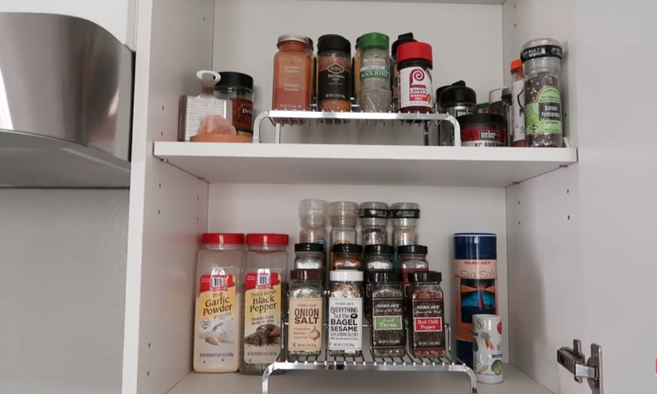 A white kitchen cabinet shelf organized with various spice jars, including large containers of garlic powder and black pepper on the bottom shelf, and smaller spice bottles on two-tiered metal risers on both the top and bottom shelves.