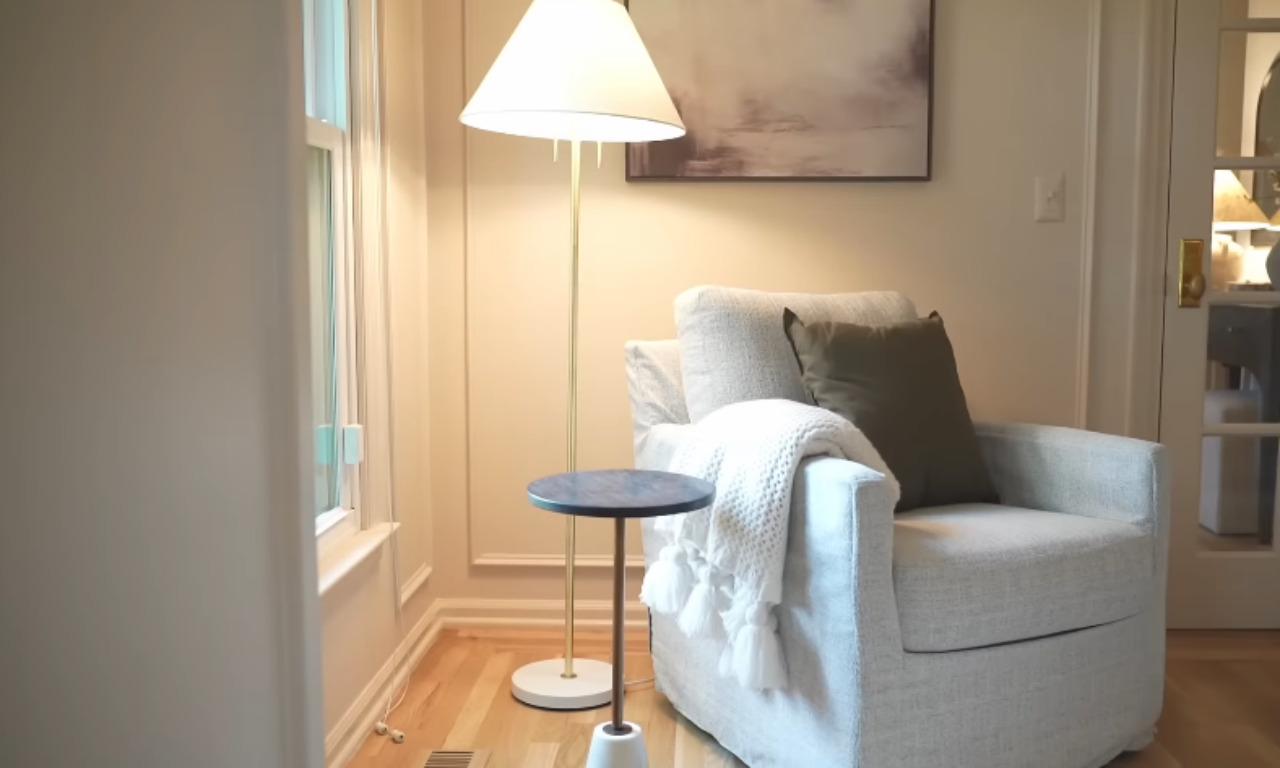 A cozy reading nook featuring a light-colored, upholstered swivel armchair with a white throw blanket draped over it and a dark olive-green cushion. A modern, brass and white floor lamp stands next to the chair, and a small, round side table with a dark top is in front of it. A framed piece of abstract art hangs on the tan paneled wall.