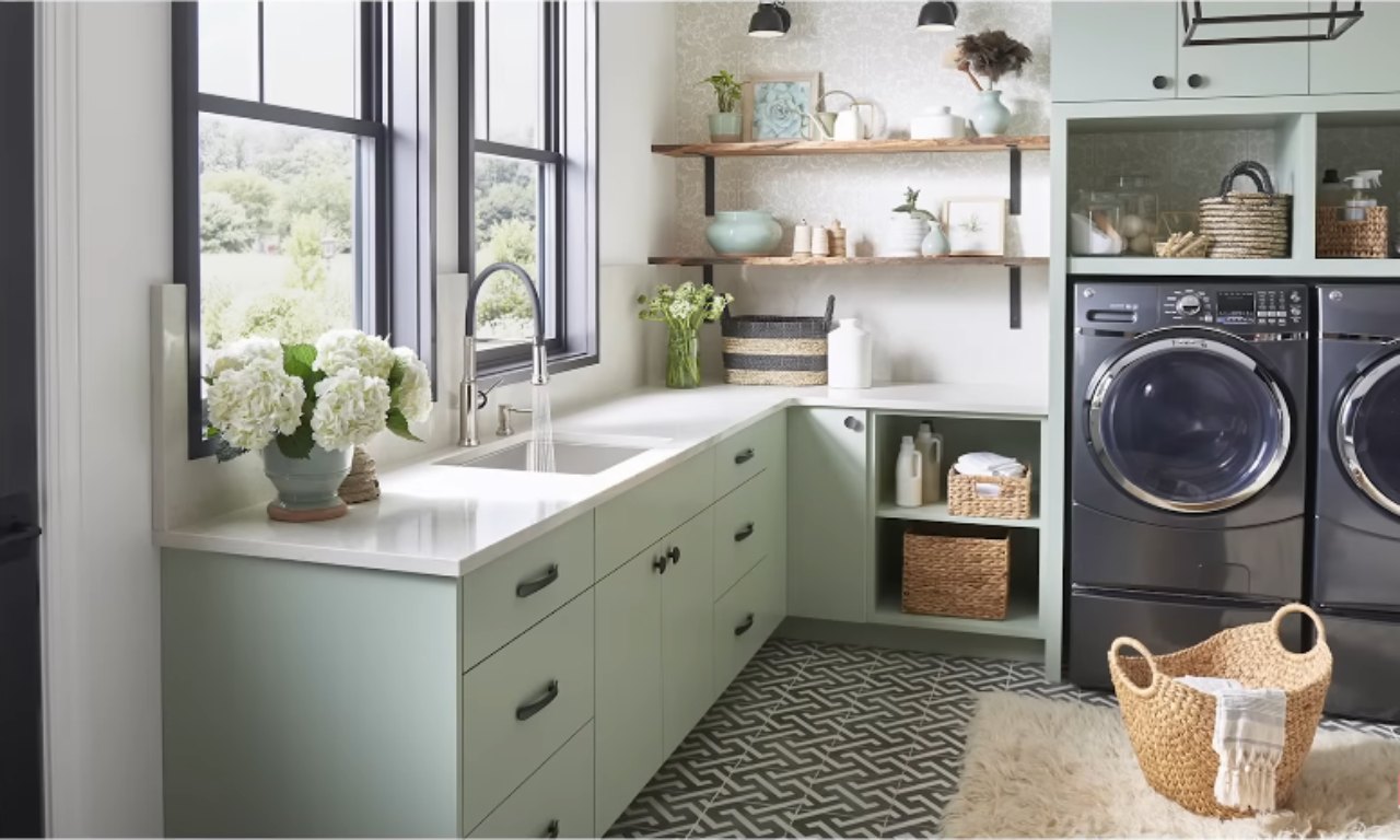 A stylish and organized laundry room with pale mint green cabinets and white countertops. Open wooden shelves above the counter hold decorative items and baskets. A stainless steel sink is by the large window. To the right, a dark gray washer and dryer are built into a cabinet unit, and a woven laundry basket sits on a geometric patterned floor.