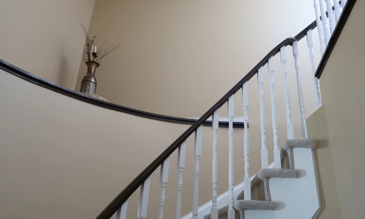 An upward-angled view of the curved staircase railing. The wall is painted a neutral beige. The dark wood handrail contrasts sharply with the white spindles and the beige-carpeted stairs. A decorative vase sits on a ledge overlooking the stairs.