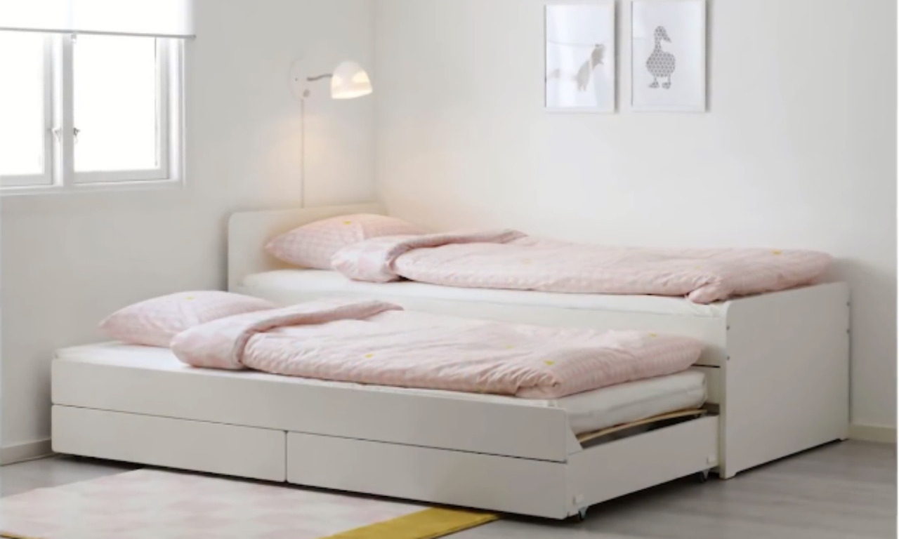 A bright, minimalist bedroom featuring a white, stacked trundle bed setup. The bottom bed is pulled out, showing two single beds side-by-side, each made with light pink and yellow patterned duvets and pillows. The walls are white, with a small window on the left and a wall-mounted reading lamp above the top bed. Two framed, simple line drawings of ducks or geese are on the wall above the top bed. A small pink and yellow checkered rug is on the floor in the foreground.