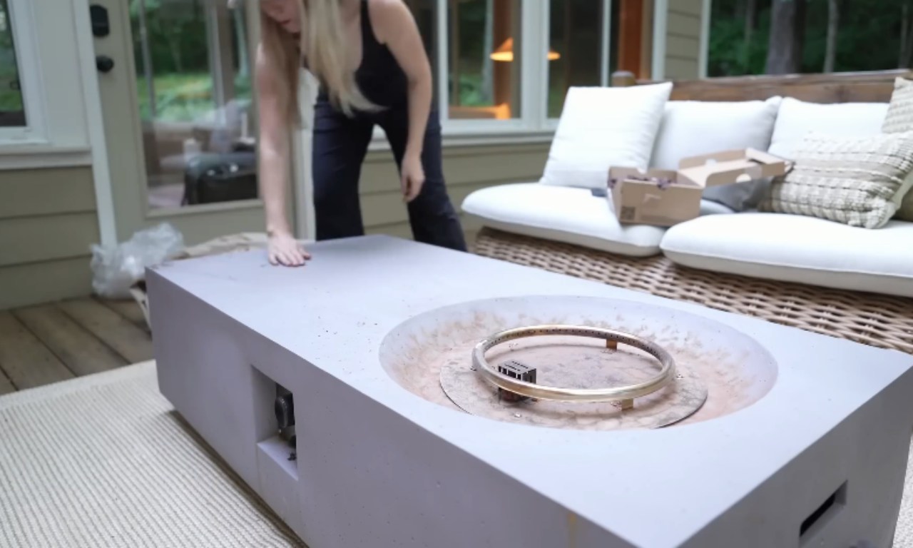 A woman in dark clothing adjusts a large, rectangular, light gray concrete fire pit table on a woven rug on an outdoor patio. A white wicker outdoor sofa with thick white cushions and pillows is visible behind the table, along with large windows looking into a home.