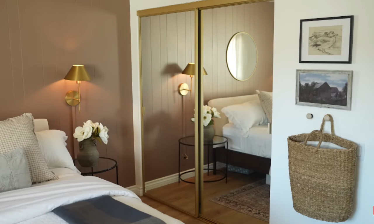 A cozy bedroom featuring a bed with white bedding, a gold-framed sliding mirrored closet door reflecting a brass wall sconce, and a gallery wall on the adjacent white wall with two framed pictures and a hanging woven basket. The bedroom wall is painted a dusty rose or mauve color.