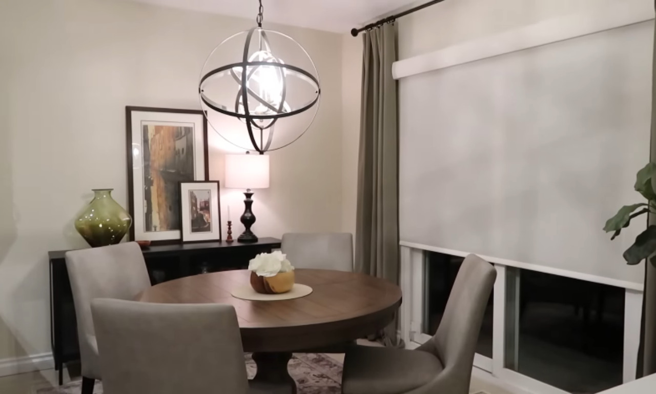 A view of a contemporary dining room. The room features a round wooden dining table surrounded by four upholstered, light gray chairs. Above the table hangs a modern, large spherical chandelier with a black metal frame. A black buffet cabinet is against the back wall, holding a large olive green vase and two framed art pieces. The room has neutral-colored walls, sheer window treatments, and long olive green curtains.