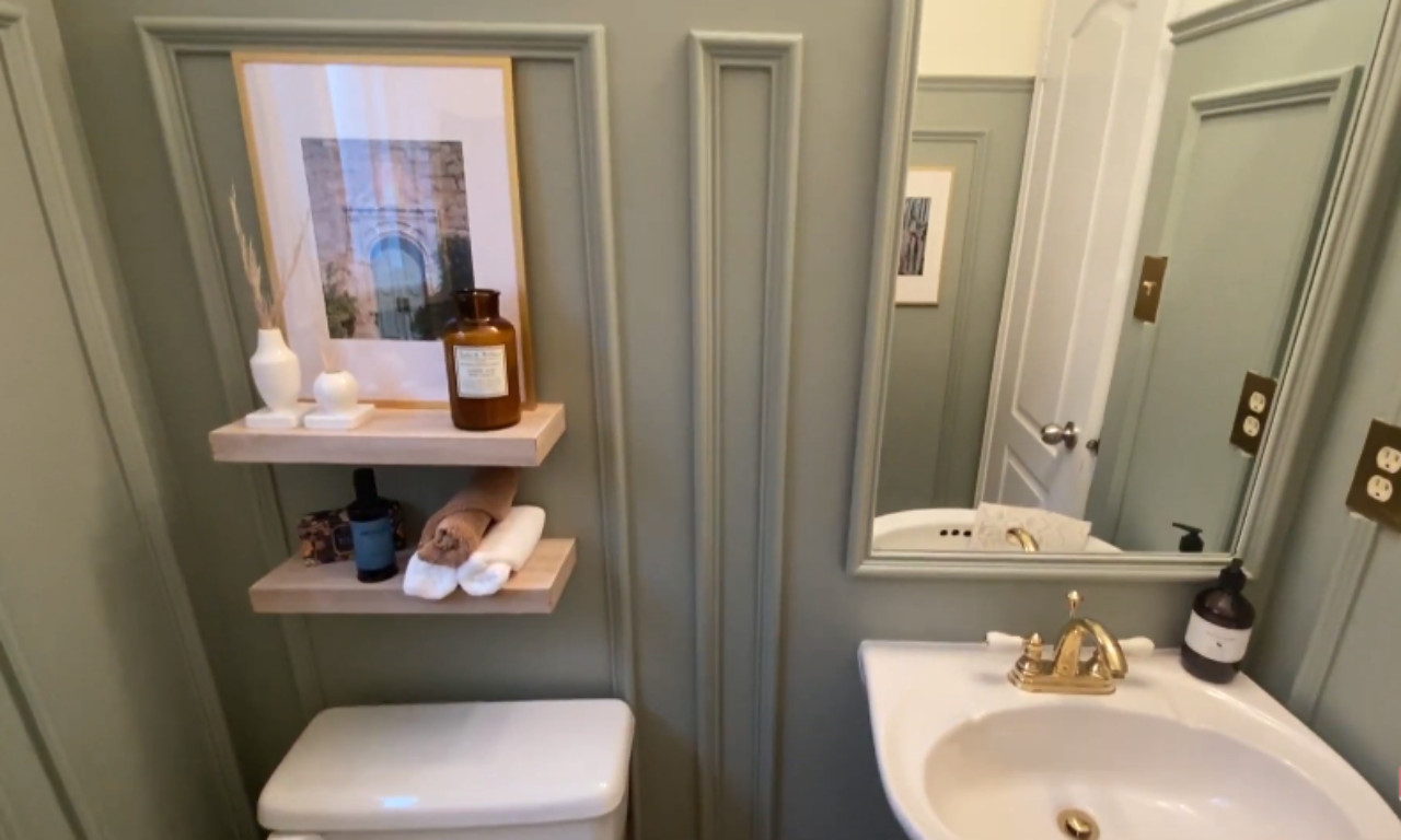 A view of a small bathroom with sage green decorative wall molding. Floating wooden shelves above a white toilet hold toiletries and small decorative items. To the right is a small white pedestal sink with a gold faucet and a rectangular mirror above it, reflecting the opposite wall.