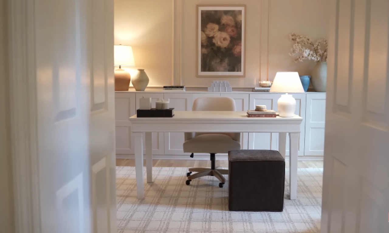 View of a calm, neutral-toned home office through an open doorway. A white desk with a beige ergonomic chair is centered on a light-colored plaid rug. White built-in cabinets line the back wall, topped with lamps, books, and decorative vases.
