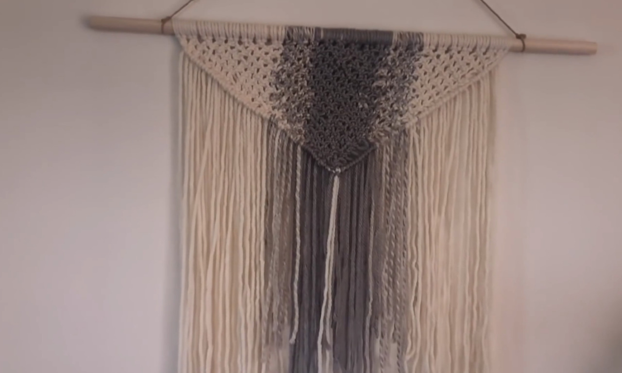 A finished DIY macrame wall hanging on a light wall. It features a woven, inverted triangle pattern in cream and gray macrame cord at the top, hanging from a wooden dowel. Long strands of cream and gray yarn/cord hang below the woven section.