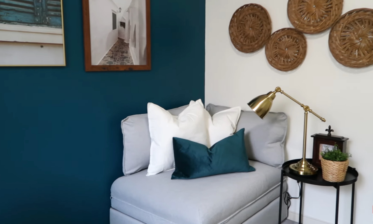 A close-up shot of a cozy seating area in a room. A grey upholstered chaise or chair is decorated with white and dark teal pillows. The background features a deep teal-colored wall to the left, and a white wall to the right, which is decorated with three circular woven rattan wall hangings. A brass-toned desk lamp rests on a small black side table next to the chair, along with a framed photo and a small potted plant in a wicker basket.
