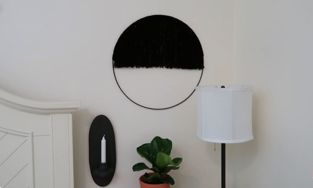 A modern, monochromatic yarn wall hanging in a bedroom setting. The piece is a large black circle or hoop, half-filled with straight-hanging black yarn fringe at the bottom. It's mounted on a white wall above a white headboard, next to a black oval sconce with a candle and a floor lamp.