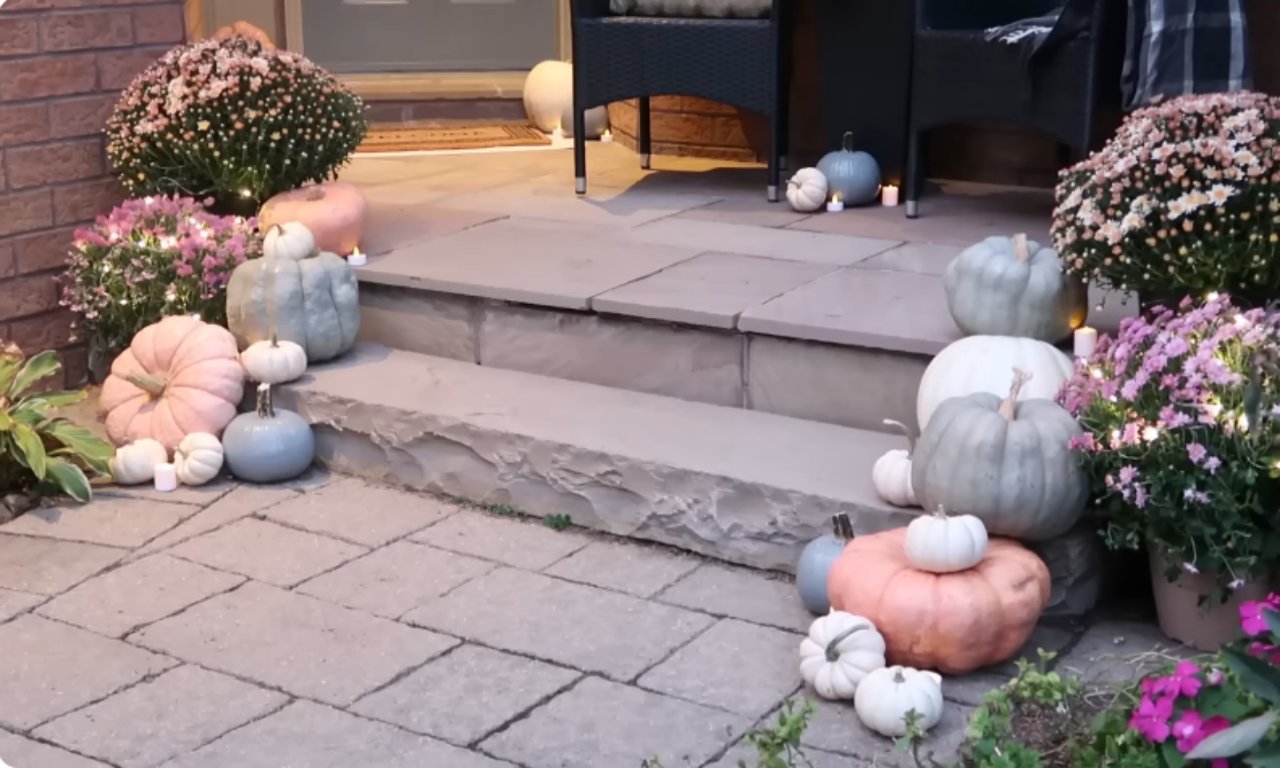 A cozy fall front porch decorated with an assortment of real pumpkins and gourds in pastel colors (dusty pinks, sage greens, and whites). Large potted chrysanthemum plants with pink and white blooms, illuminated by small twinkle lights, flank the stone steps. Small white tea lights or candles are interspersed among the pumpkins on the patio paving and steps.