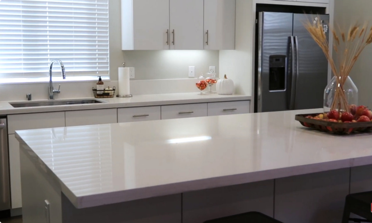 A bright, modern kitchen with white cabinets, light gray walls, and white countertops. A central kitchen island with a light countertop is in the foreground. Countertop decor includes a vase of dried wheat stalks, small pumpkins, and a tray filled with red apples, suggesting a fall theme.
