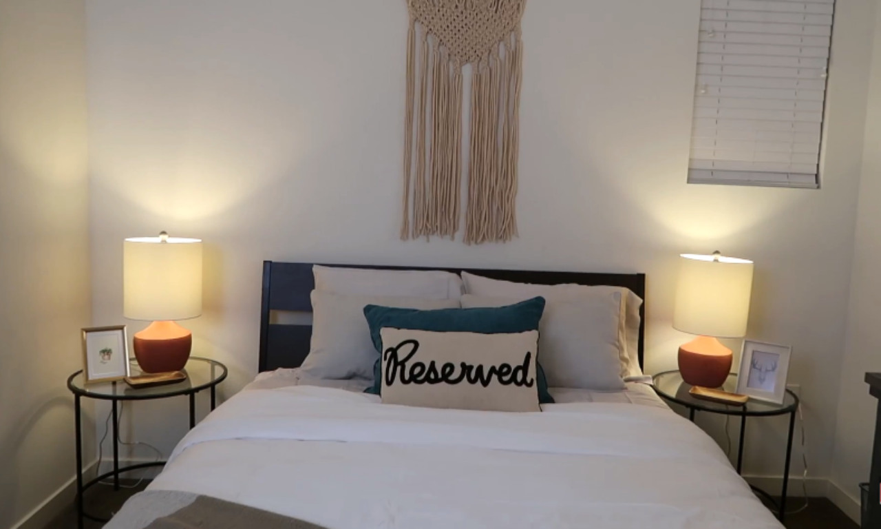A view of a well-decorated guest room with a full or queen-size bed centered against a white wall. The bed has white bedding and is accented with two pillows: a solid teal one and a decorative rectangular pillow that says "Reserved." A macrame wall hanging is above the headboard. On either side of the bed, a black metal side table holds a lamp with a terracotta-colored base and a warm-toned shade, along with small framed photos.