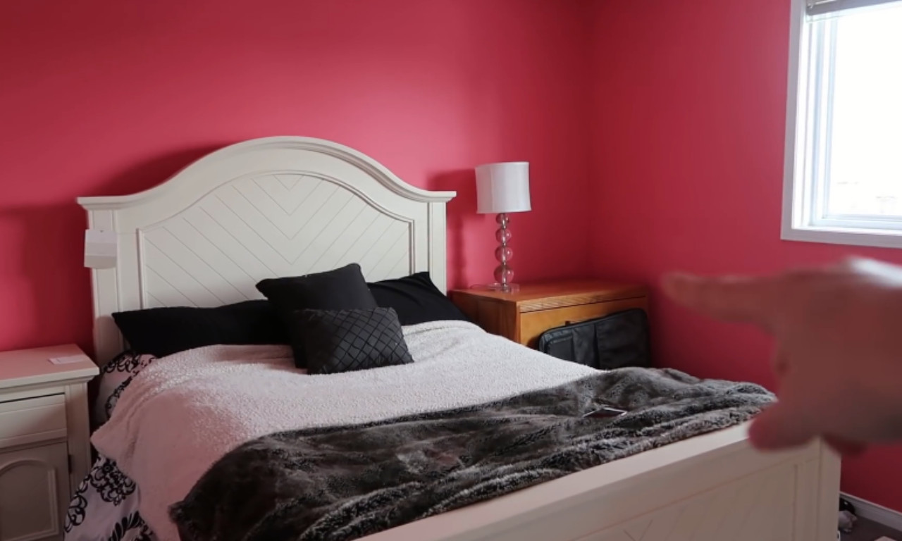 A bedroom is shown with bright magenta-pink walls. The room contains a white headboard on a bed with a white, fluffy comforter and black and dark gray accent pillows and a throw blanket. A wooden nightstand is to the right, and a window is visible in the corner. A person's blurred finger points toward the right side of the room.