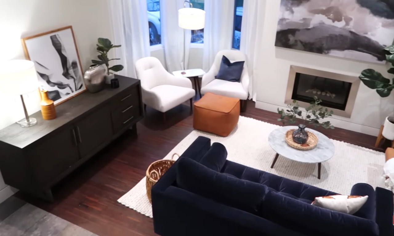 Overhead view of a modern living room with dark wood floors and a light area rug. A navy blue velvet sofa is in the foreground, facing a round marble coffee table. In the background, there is a dark wooden console table with decor, two white accent chairs, a brown leather ottoman, and a fireplace with abstract art hanging above it.