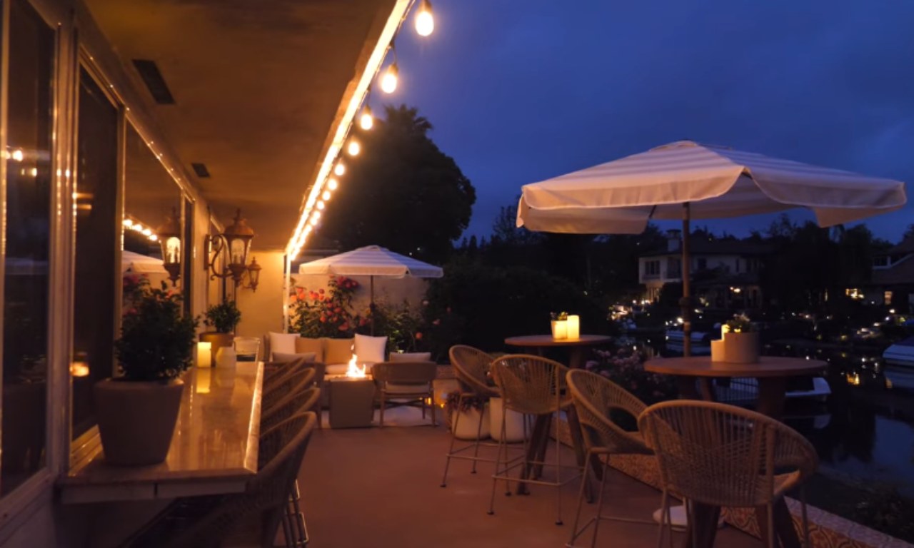 A patio at dusk or night, illuminated by string lights running along a covered structure. The scene includes a long bar area on the left with glowing candles and potted plants. On the right, there are various seating zones with round tables, wicker bar stools, a small fire pit, and a striped umbrella. The background is dark with lights reflecting on the water and from the distant houses.
